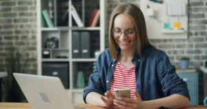 Competitive Compensation: a woman sitting at a table using smartphone at work swiping looking at screen and smiling